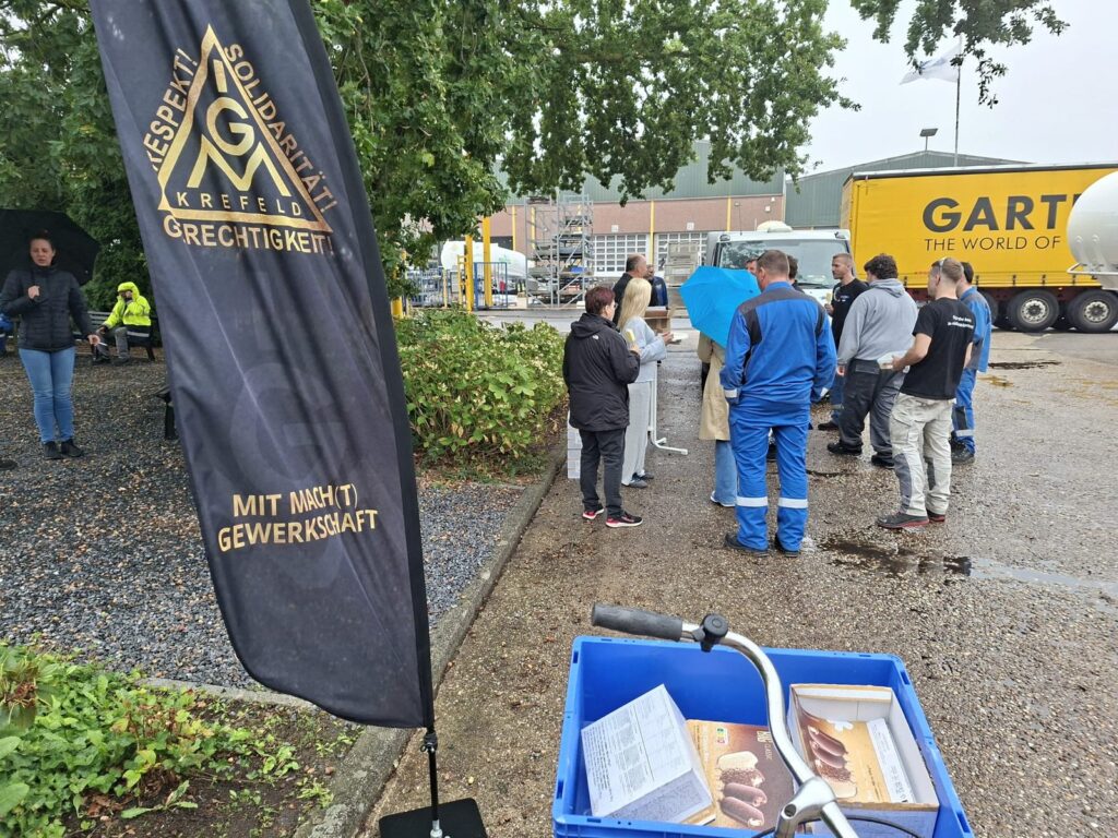 Beachflag der IG Metall Krefeld im Vordergrund, dahinter Kolleginnen und Kollegen von GOFA mit Eis im Innenhof